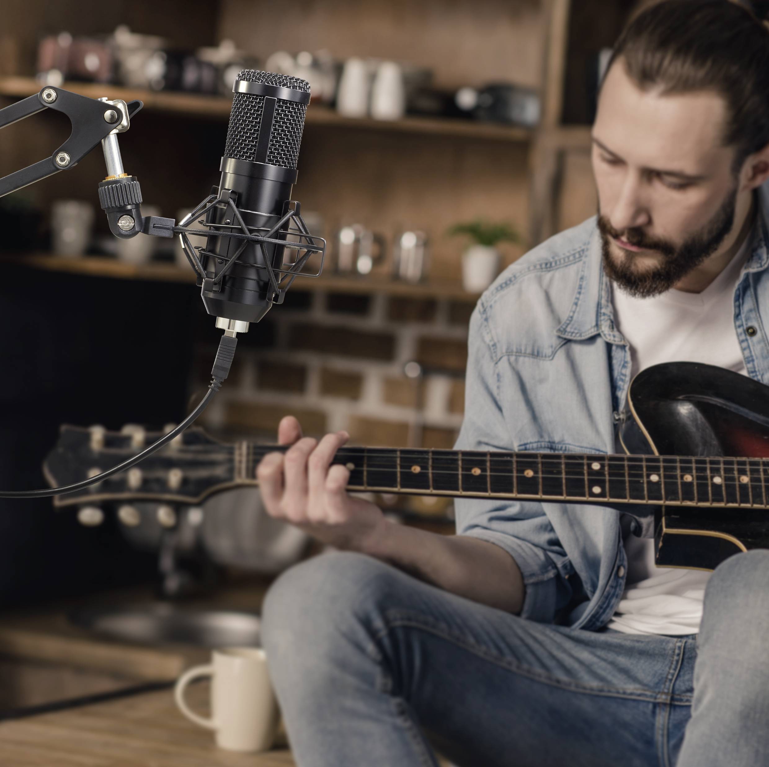 En man spelar gitarr i ett mysigt rum. En mikrofon är riktad mot honom, sannolikt för en inspelning eller streaming.