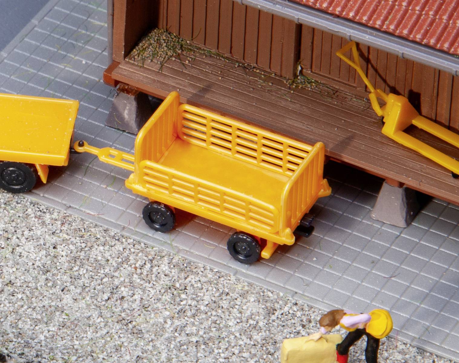 En gul trailer står bredvid en byggnad med rött tak. En arbetare bär en låda. Scenen ser ut som en miniatyrmodell.