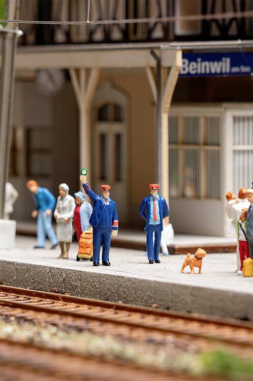 En miniatyrmodell av ett tågstation med människor och en hund på perrongen. En konduktör höjer armen, en skylt visar 'Beinwil'.