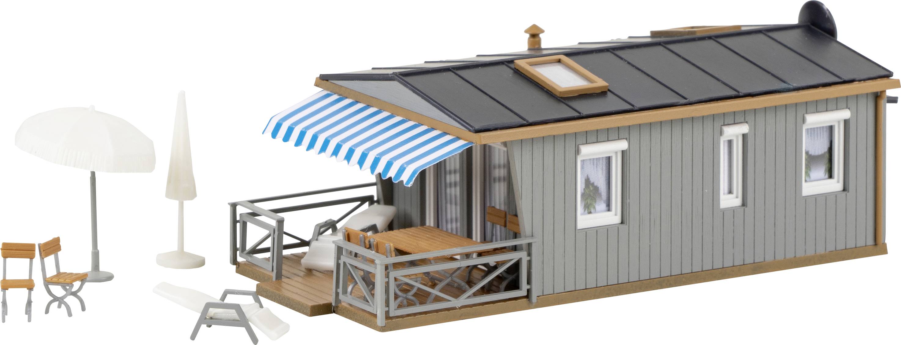 En grå modelltillverkad husvagn med blå-vit randig markis, liten veranda, bord, stolar, parasoll och solstol.