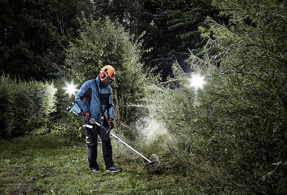 En person i arbetsklädsel med hjälm och hörselskydd använder en röjsåg för att klippa högt gräs och buskar i ett skogsområde.