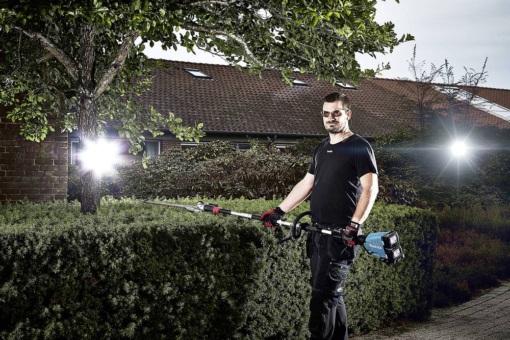 En man i arbetsklädsel använder en elektrisk häcksax för att klippa en häck. I bakgrunden syns träd och ett hustak.