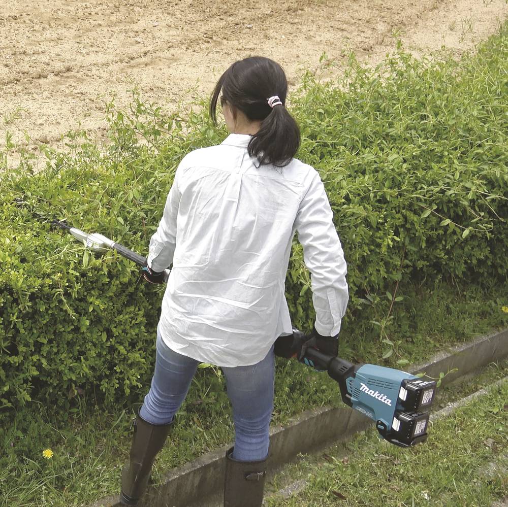 En person klipper en häck med en sladdlös häcksax på en gräsmatta. Personen bär gummistövlar och en jacka.