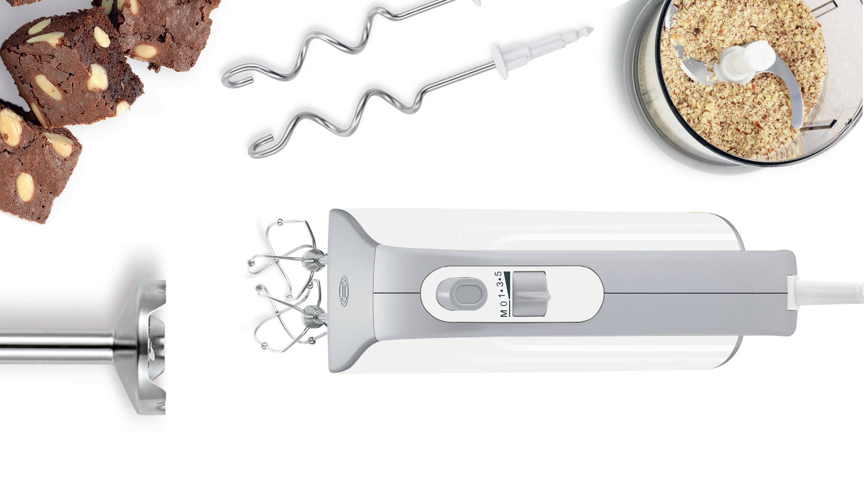 Elektrisk handmixer med olika tillbehör, däribland visp och degkrok. Bredvid finns en skål med malda nötter och chokladkaka.