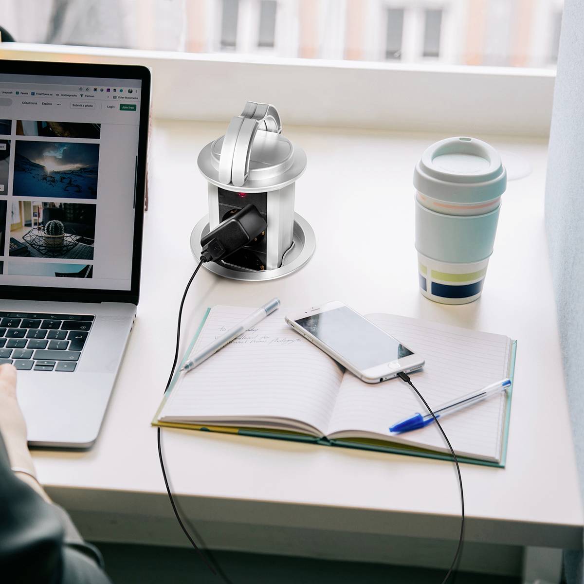 En bärbar dator visar en e-handelswebbplats. Bredvid den finns en laddare med en smarttelefon och en anteckningsbok med en penna. En kopp står intill.