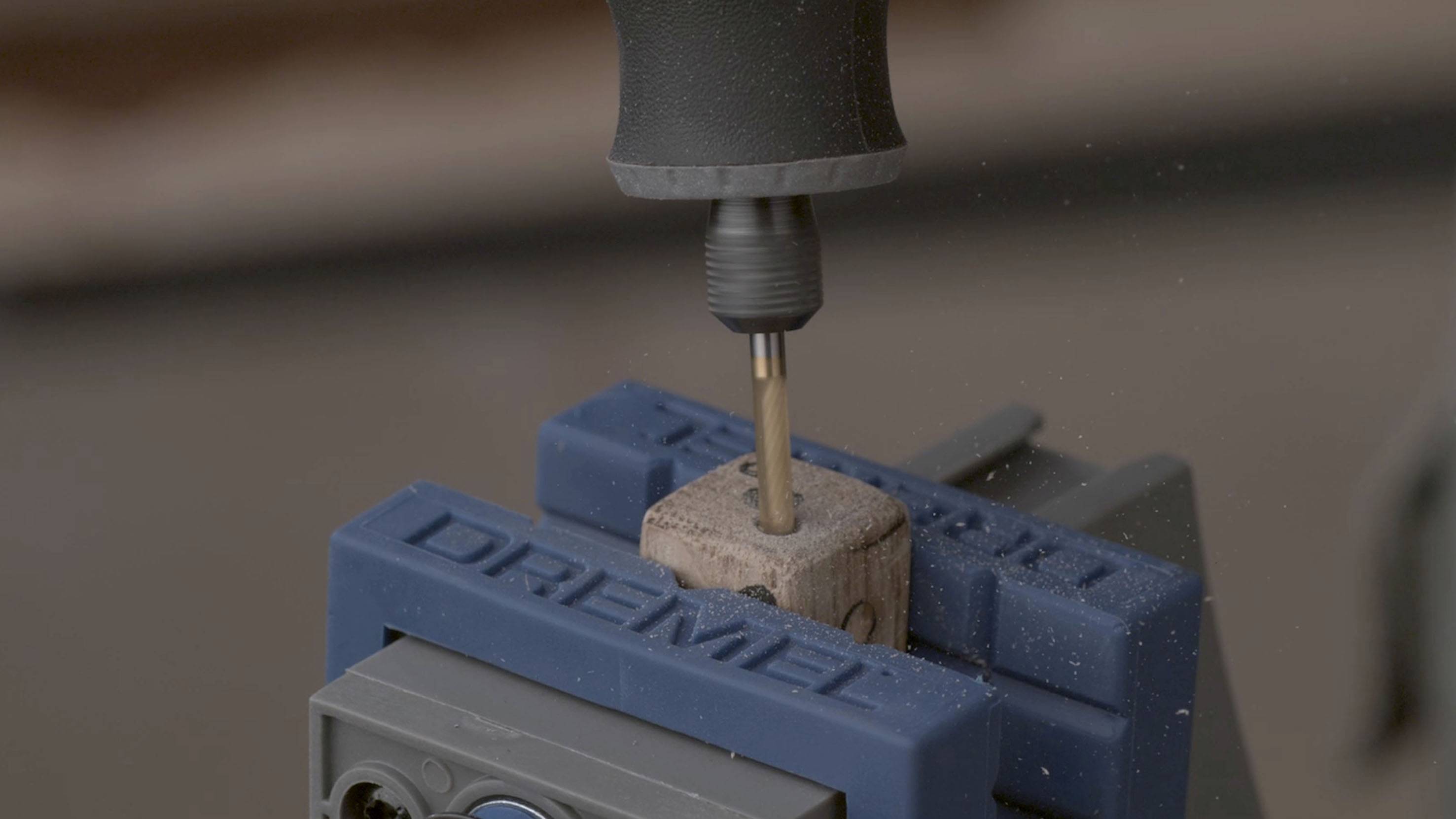 Träbit inspänt i en blå skruvstäd med ett Dremel-verktyg, medan en borr borrar ett hål.