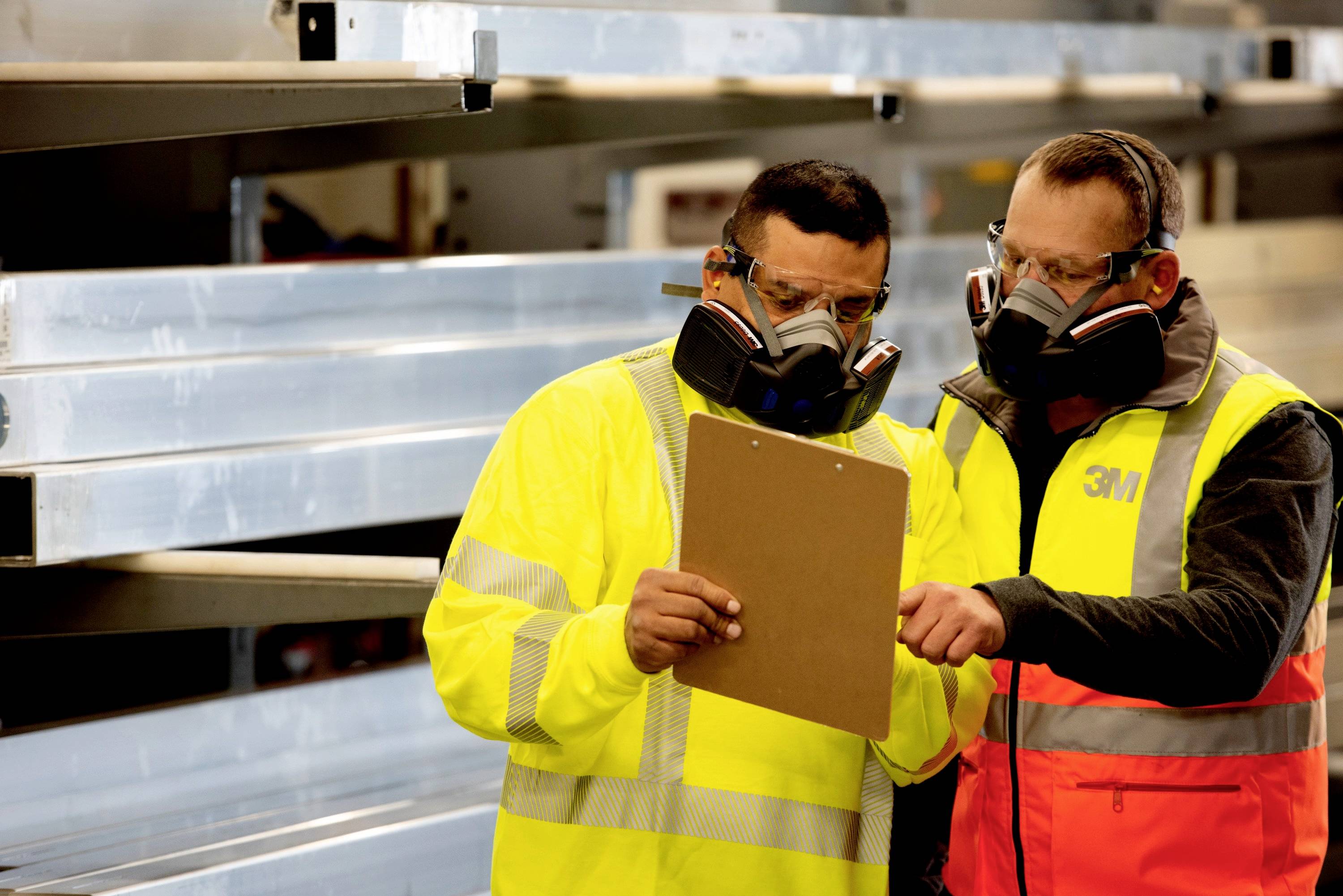 Två män i skyddskläder och andningsskydd kontrollerar en clipboard i en industriell miljö med metallhyllor i bakgrunden.