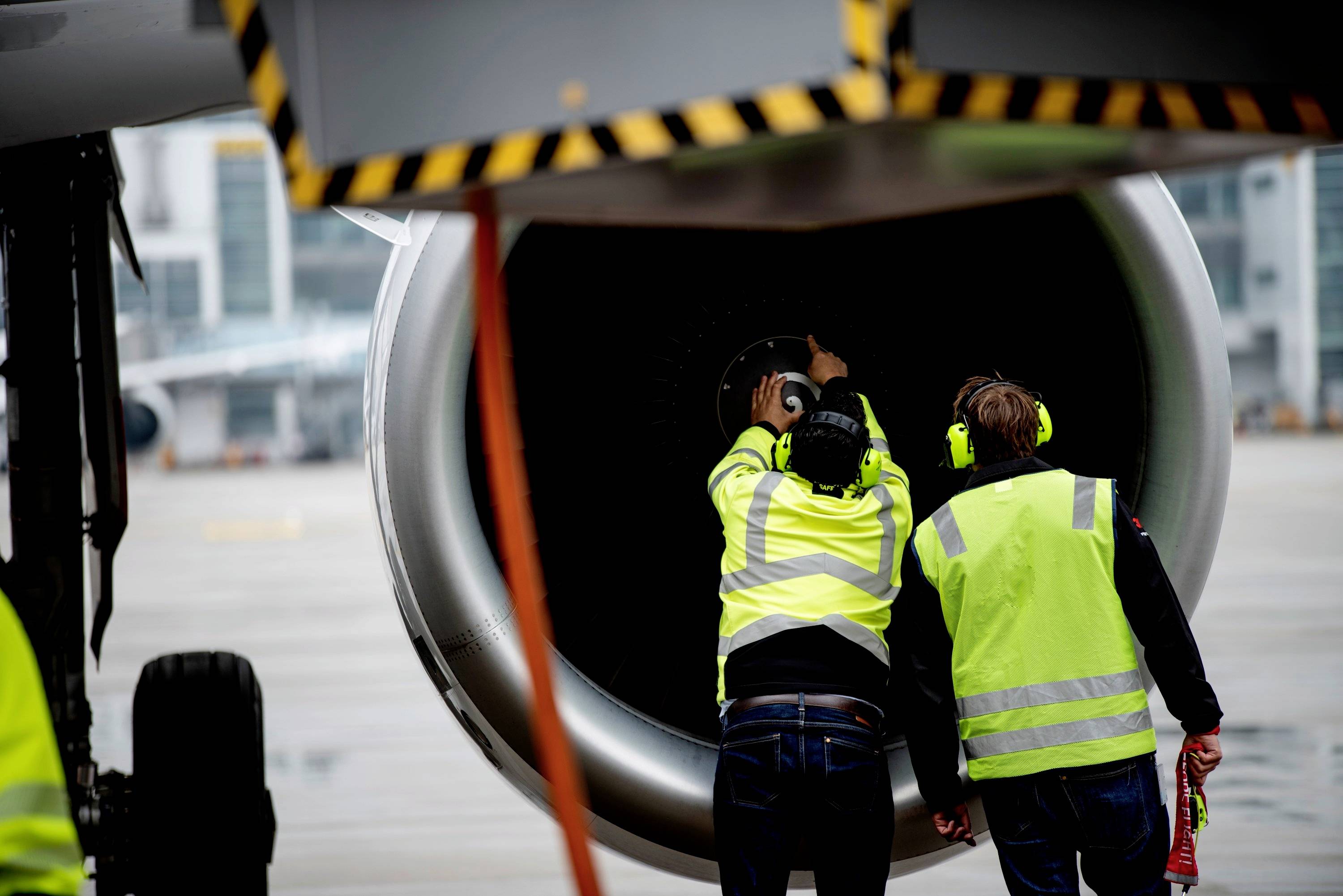 Två teknikexperter inspekterar ett flygplans motor på startbanan. De bär varselvästar och hörselskydd.