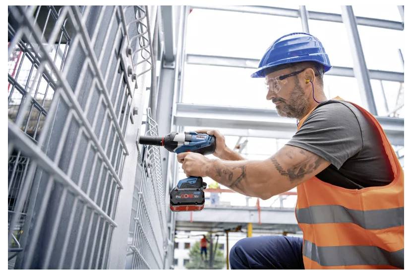 En byggnadsarbetare som bär en blå skyddshjälm och orange varselväst använder en sladdlös skruvdragare på en metallstruktur vid en byggarbetsplats.