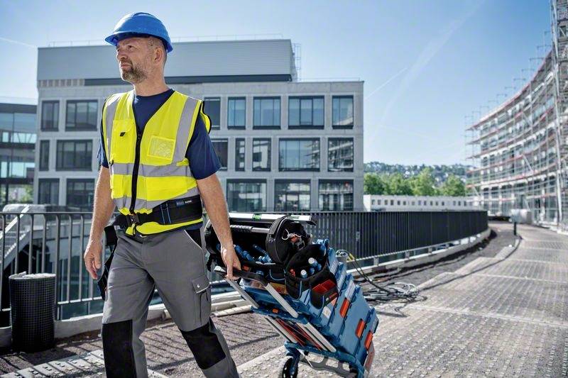 En byggnadsarbetare bär en varselväst och en hjälm. Han drar en verktygslåda på en vagn över en gångväg framför moderna kontorsbyggnader.