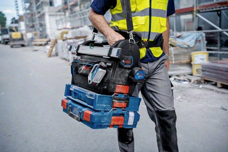 Byggnadsarbetare i skyddsväst, bär verktygslåda på byggarbetsplatsen. Bakgrunden visar byggnadsställning och högar av byggmaterial.