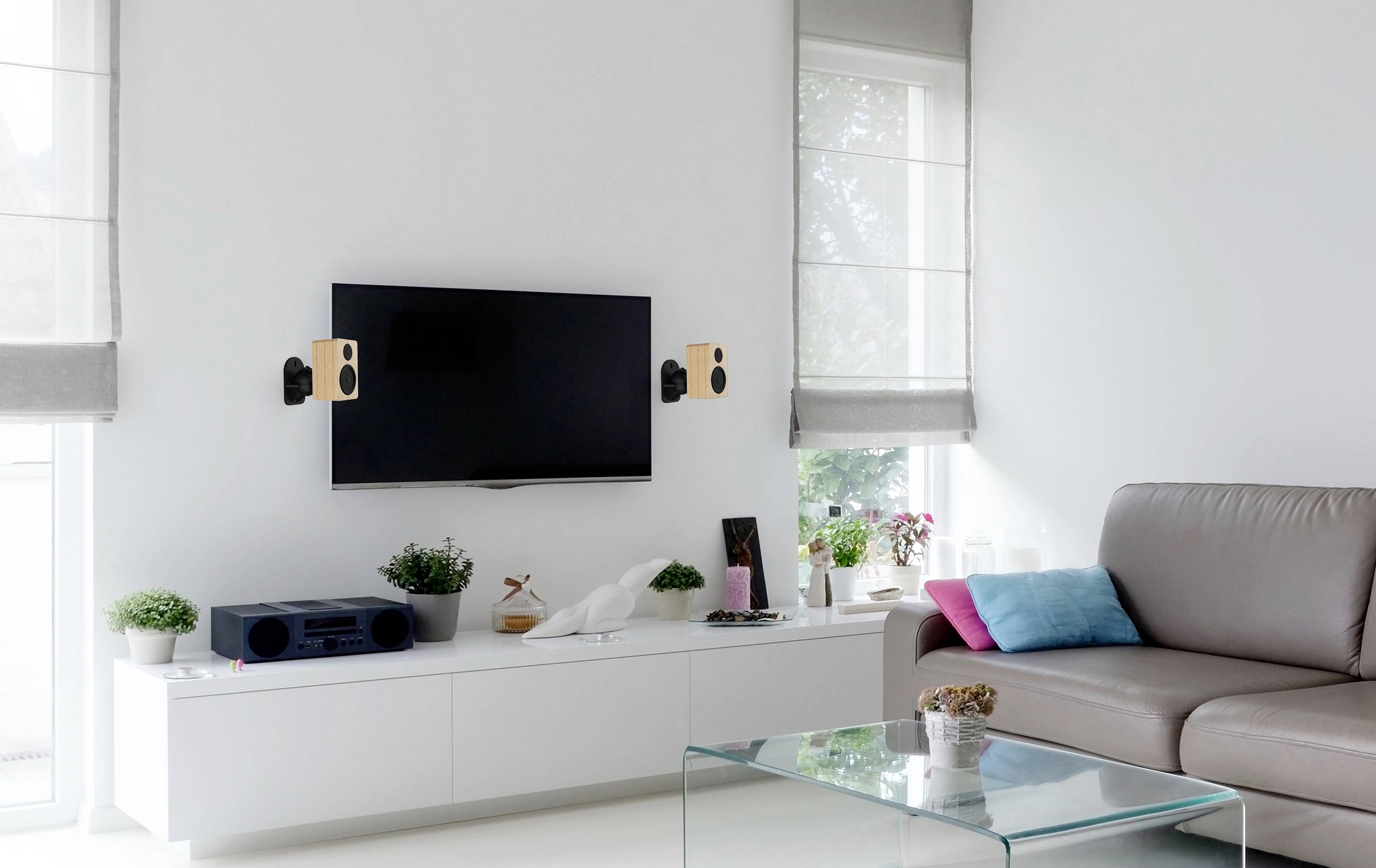 Modernt vardagsrum med vitt interiör; platt-TV på väggen, högtalare, grå soffa, dekorationer och växter på vitt sideboard.