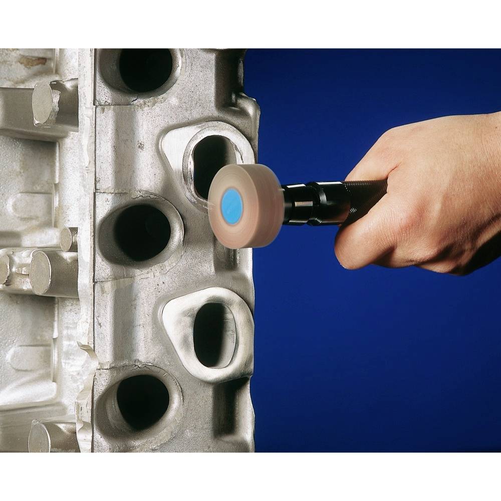 En hand håller ett verktyg som sliper ett metallhölje med olika öppningar. Scenen visar en precisionsarbetsuppgift.