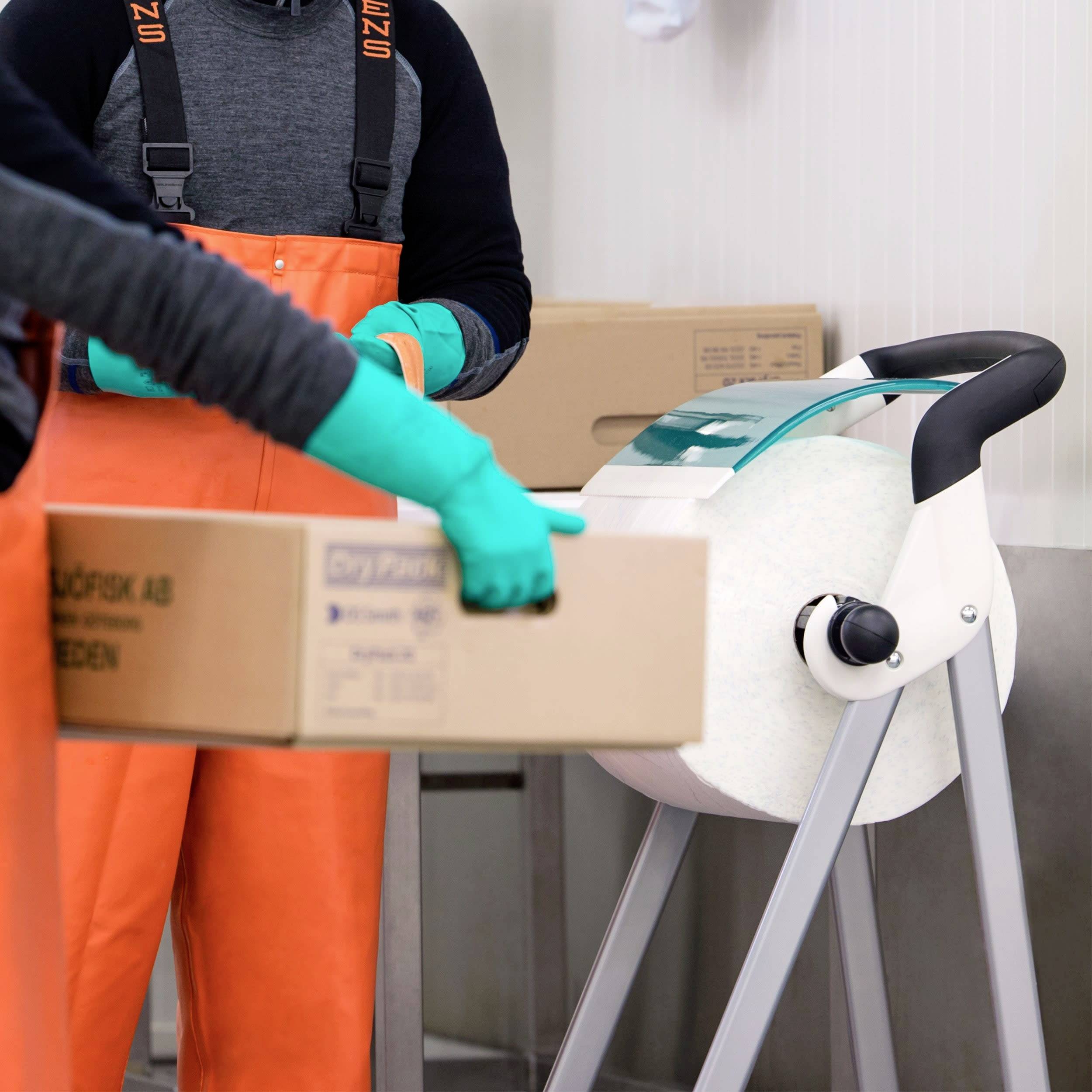 Två personer i orange arbetskläder och gröna handskar bär lådor i ett lagerutrymme. I bakgrunden syns fler lådor.
