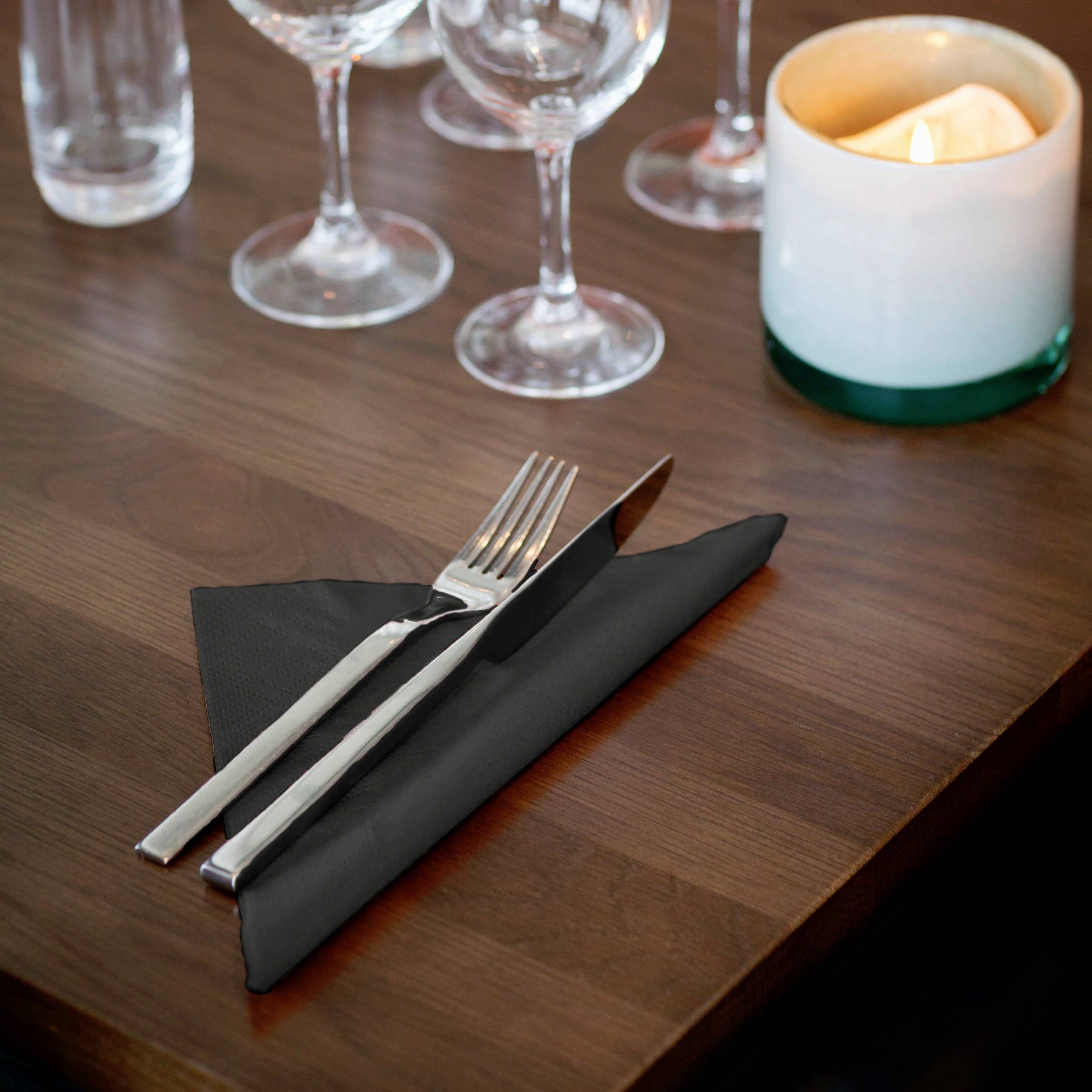 Bestick bestående av kniv och gaffel ligger på en svart tygservett på bordet. I bakgrunden brinner ett värmeljus och tomma vinglas.