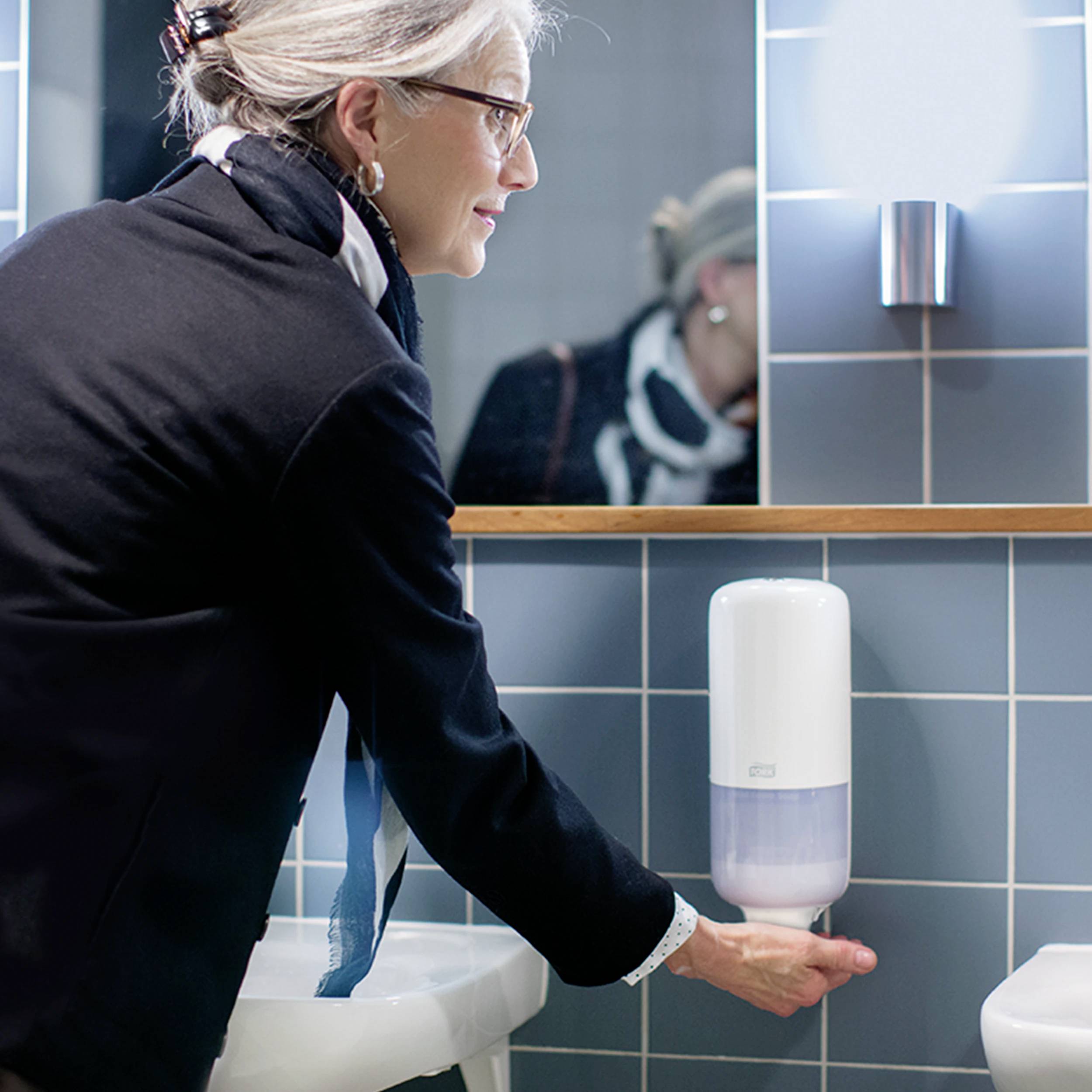 En person i ett badrum trycker på en tvålbehållare monterad på väggen. I bakgrunden syns blå kakel.