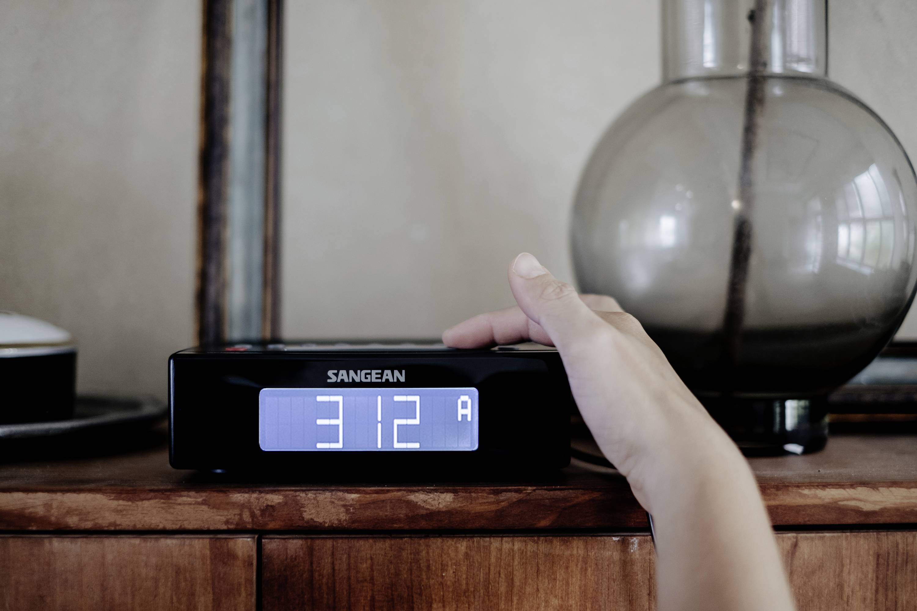 En hand ställer in tiden på en digital väckarklocka som visar '3:12 FM'. I bakgrunden står en glasvas.