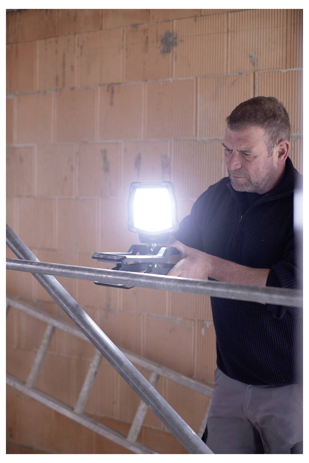 En man installerar ett starkt arbetsljus på metallställningar inuti en ofärdig tegelbygnad.