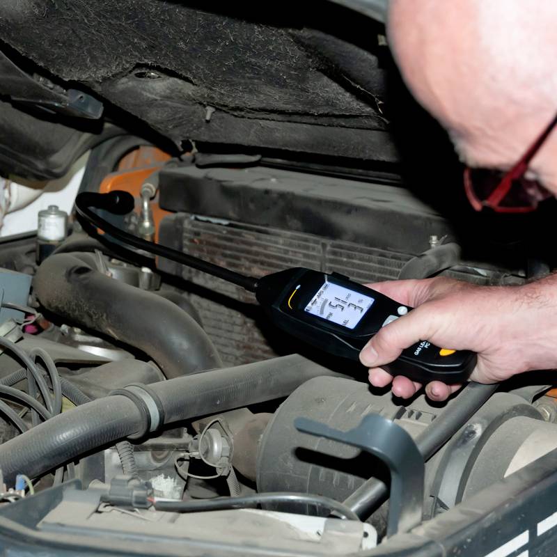En person mäter motorns temperatur med en digital termometer. Motorrummet är öppet och visar motordelar.
