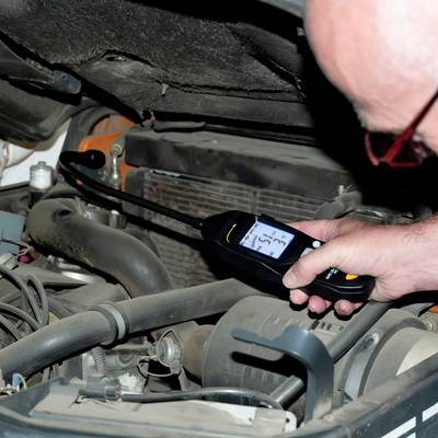 En person mäter motorns temperatur med en digital termometer. Motorrummet är öppet och visar motordelar.