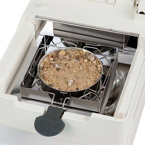 Metallbehållare med sand och stenar i en apparat, möjligen för analys av materialprover i en laboratoriemiljö.