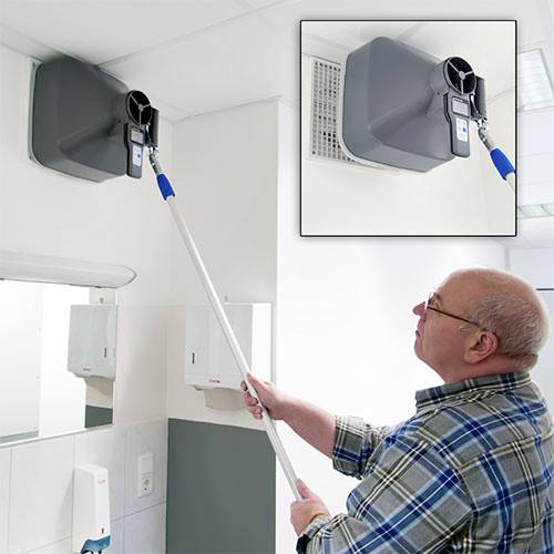 En man mäter luftflödet vid ett ventilationsintag i ett kontor eller laboratorium med hjälp av en mätenhet som är monterad i taket.