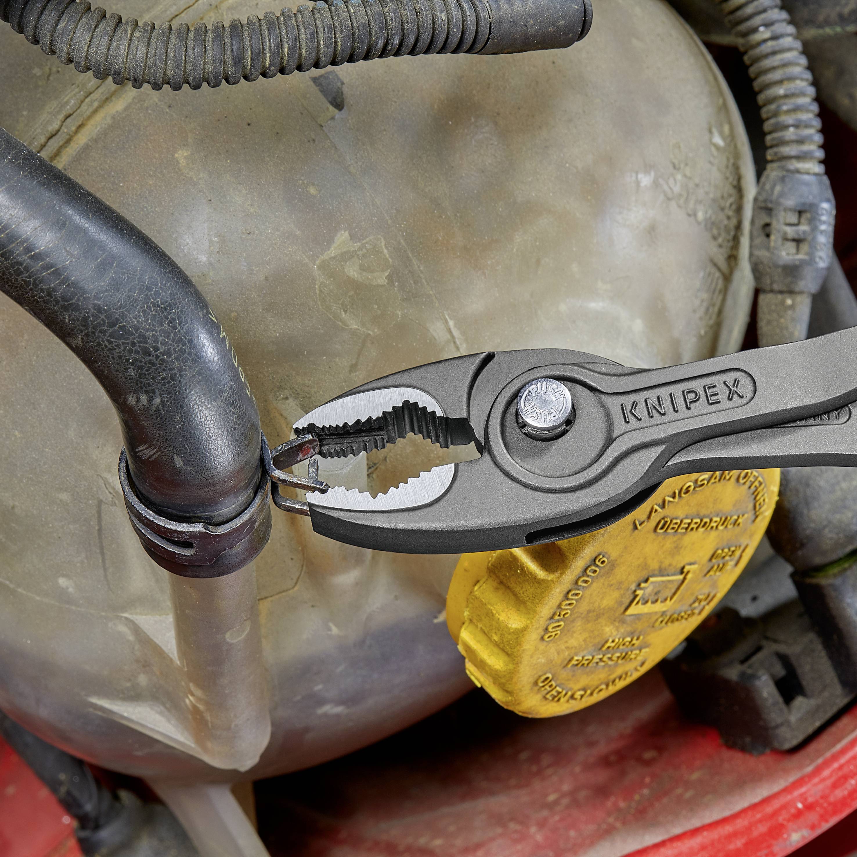 En KNIPEX-tång håller en gummislang, omgiven av metalldelar, i en verkstadsmiljö.