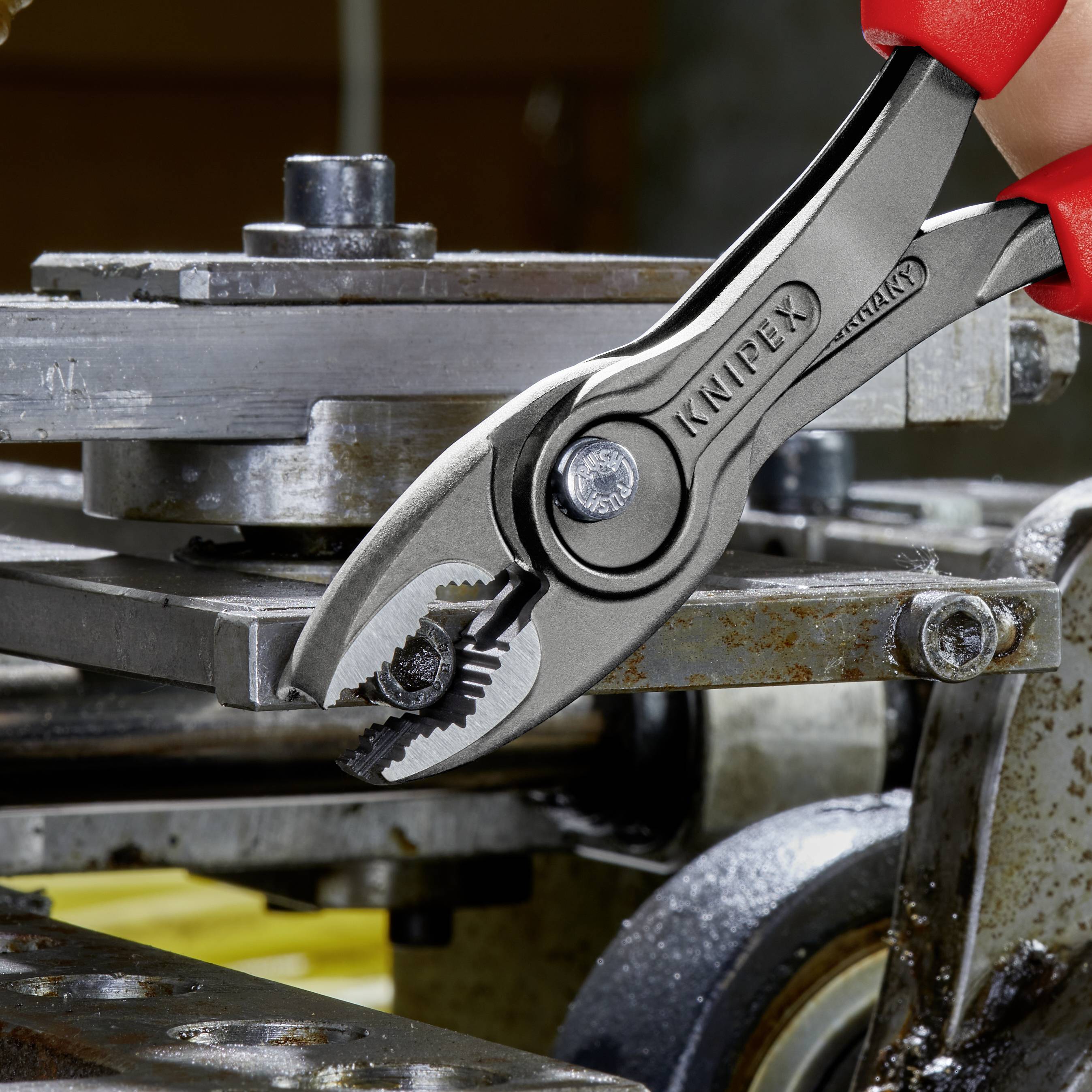 En röd hand håller en Knipex-tång som griper ett stycke metall i en verkstad. I bakgrunden syns industriella maskiner.