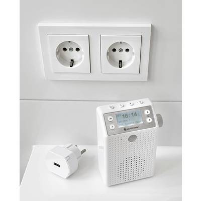 En vit FM-radio med digitalt display visar '16:14'. Bredvid ligger en adapter med två dubbla vägguttag på en vit vägg.