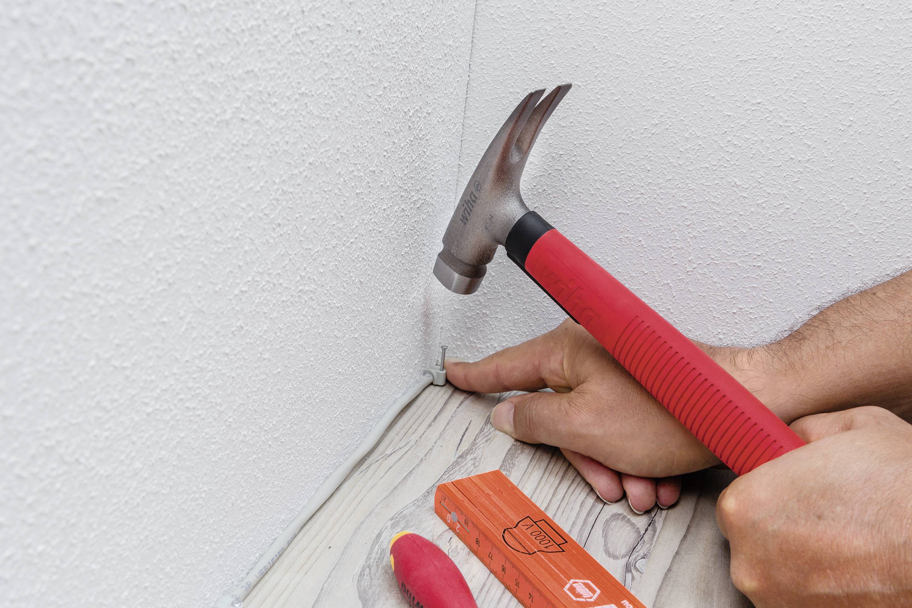 En person hamrar en spik i hörnet av en vägg för att fästa en vit kabel. På golvet ligger ett orange verktyg.