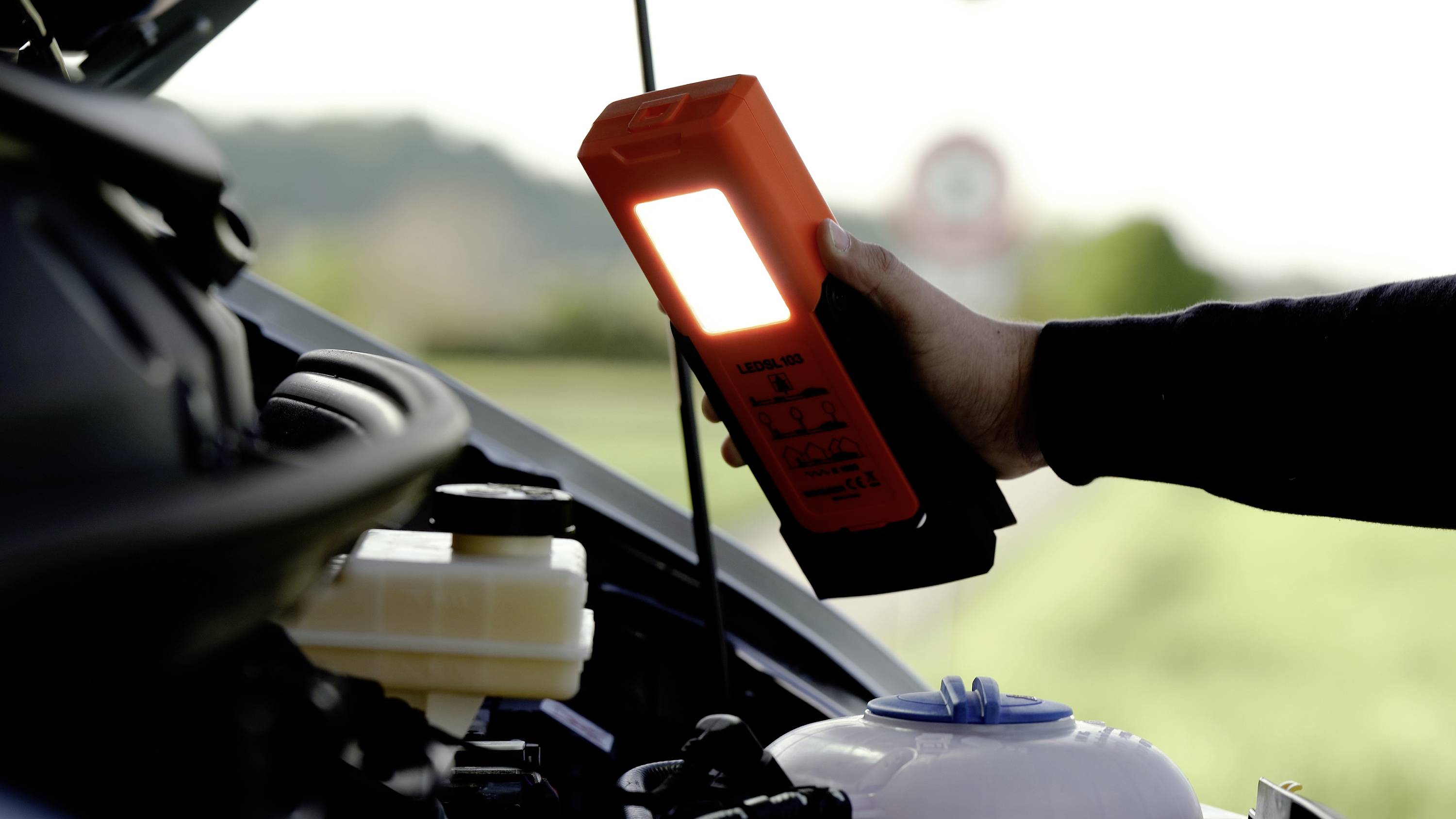 En person inspekterar en bils motor utomhus med en bärbar, lysande lampa för att förbättra sikten.