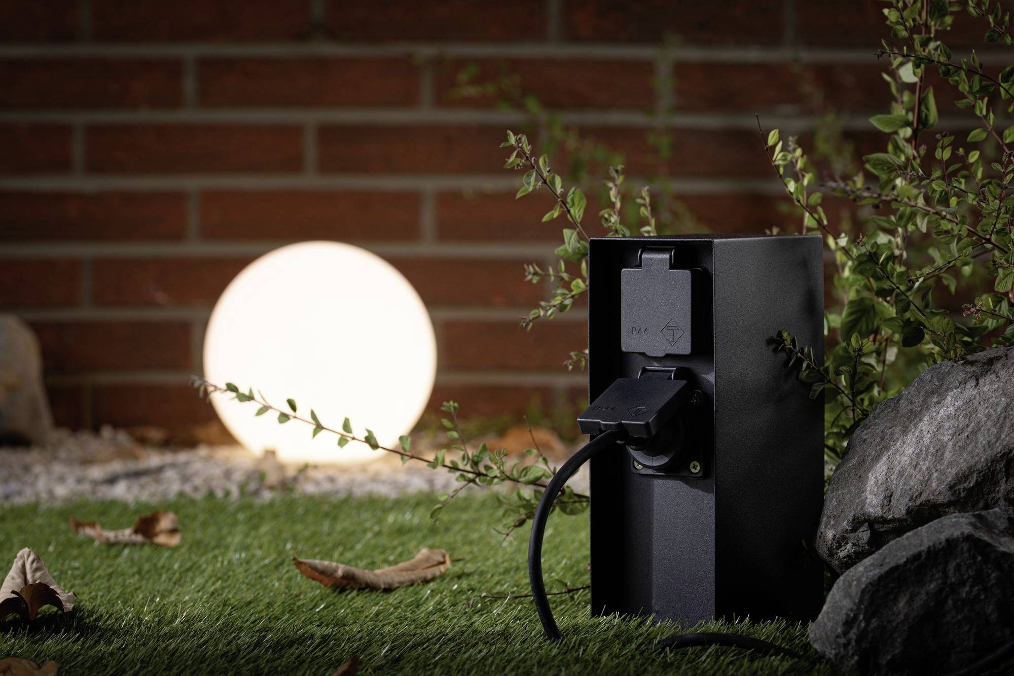 En svart kontakt står på en gräsmatta, belyst av en rund lampa i bakgrunden. Löv och en mur syns.