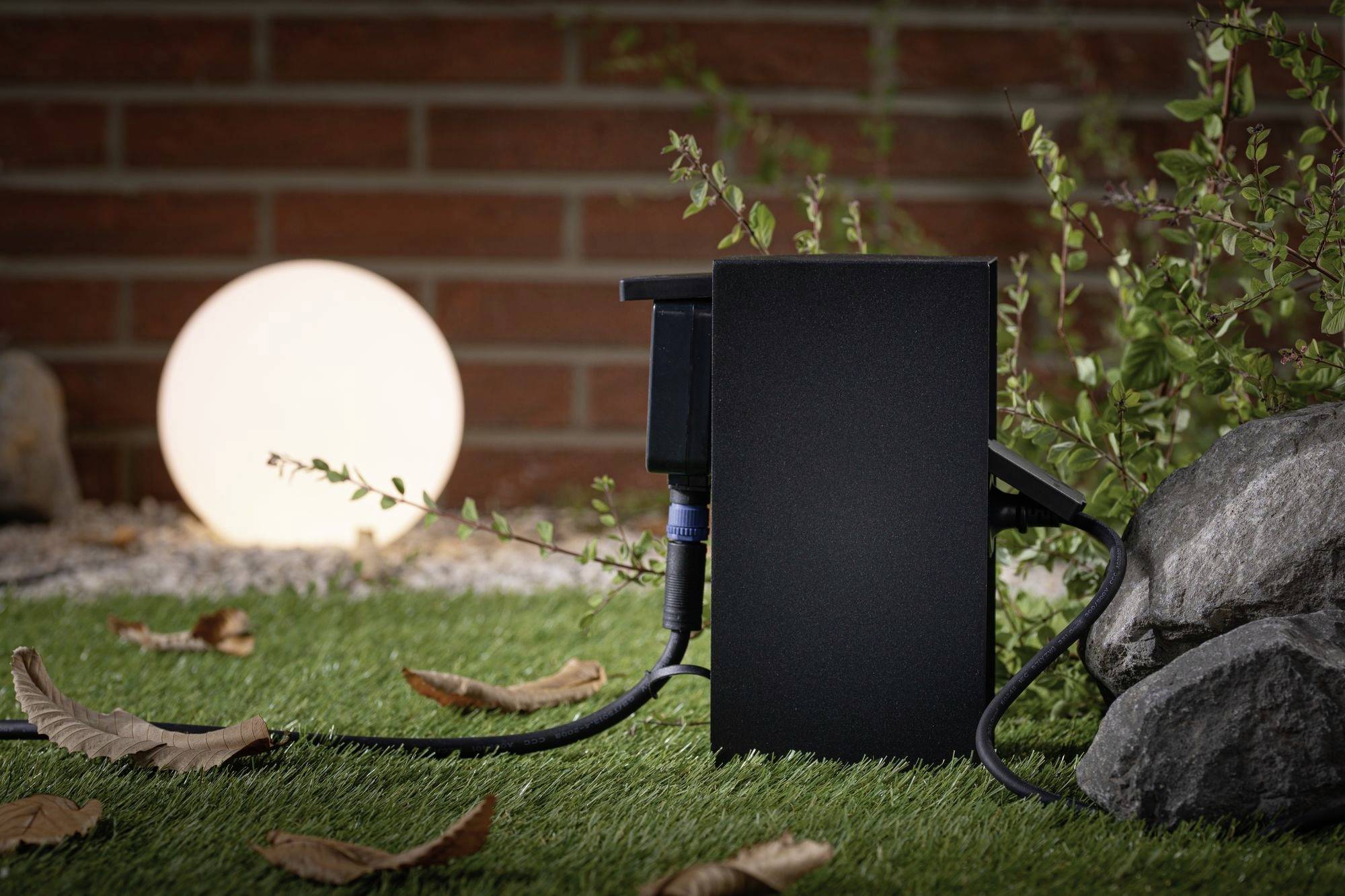 Elektrisk låda på konstgräs, omgiven av stenar, med kablar. I bakgrunden en lysande sfärisk lampa.