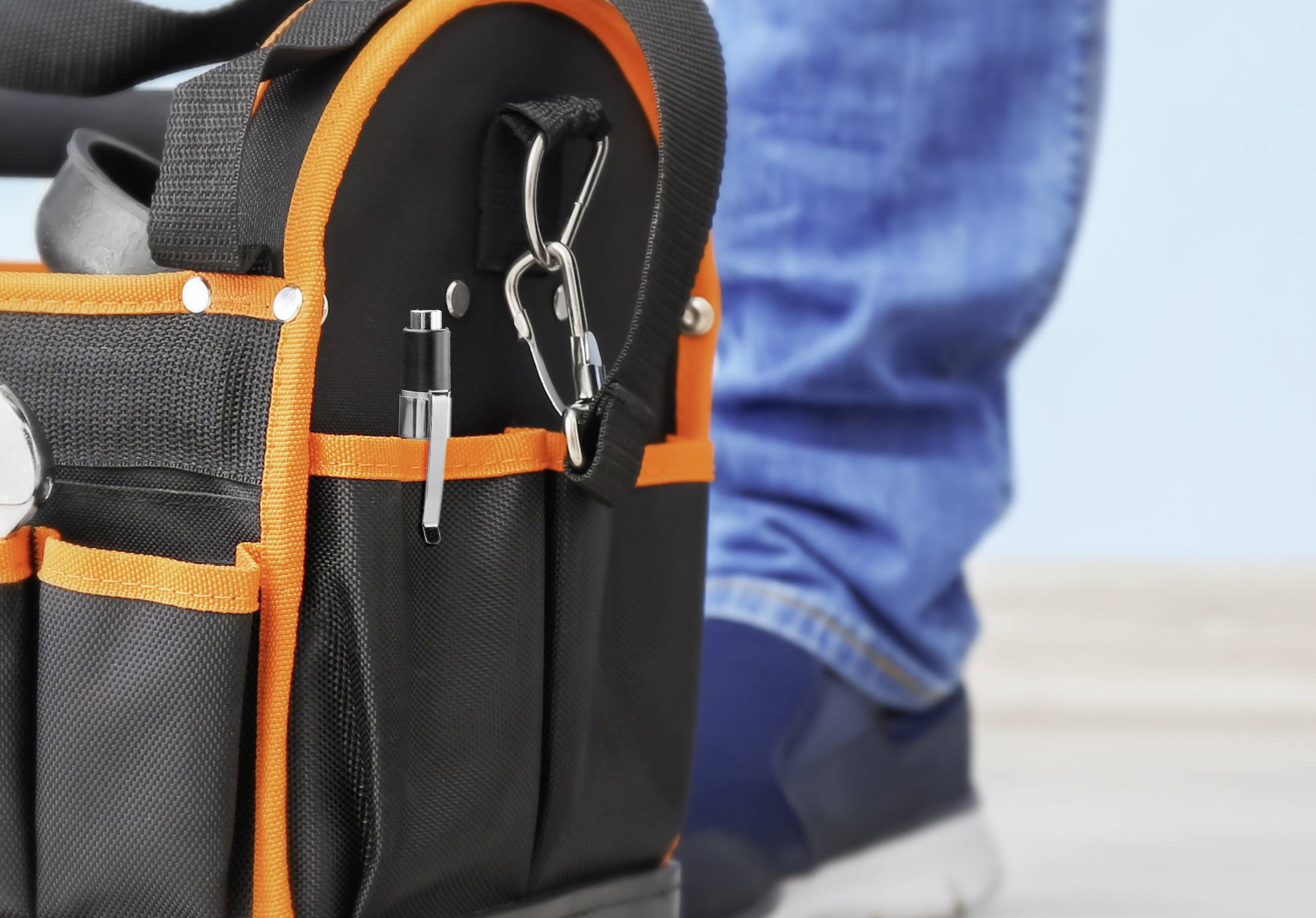 En svart verktygsväska med orange sömmar står på ett golv. En person i blå jeans och sneakers står bredvid.