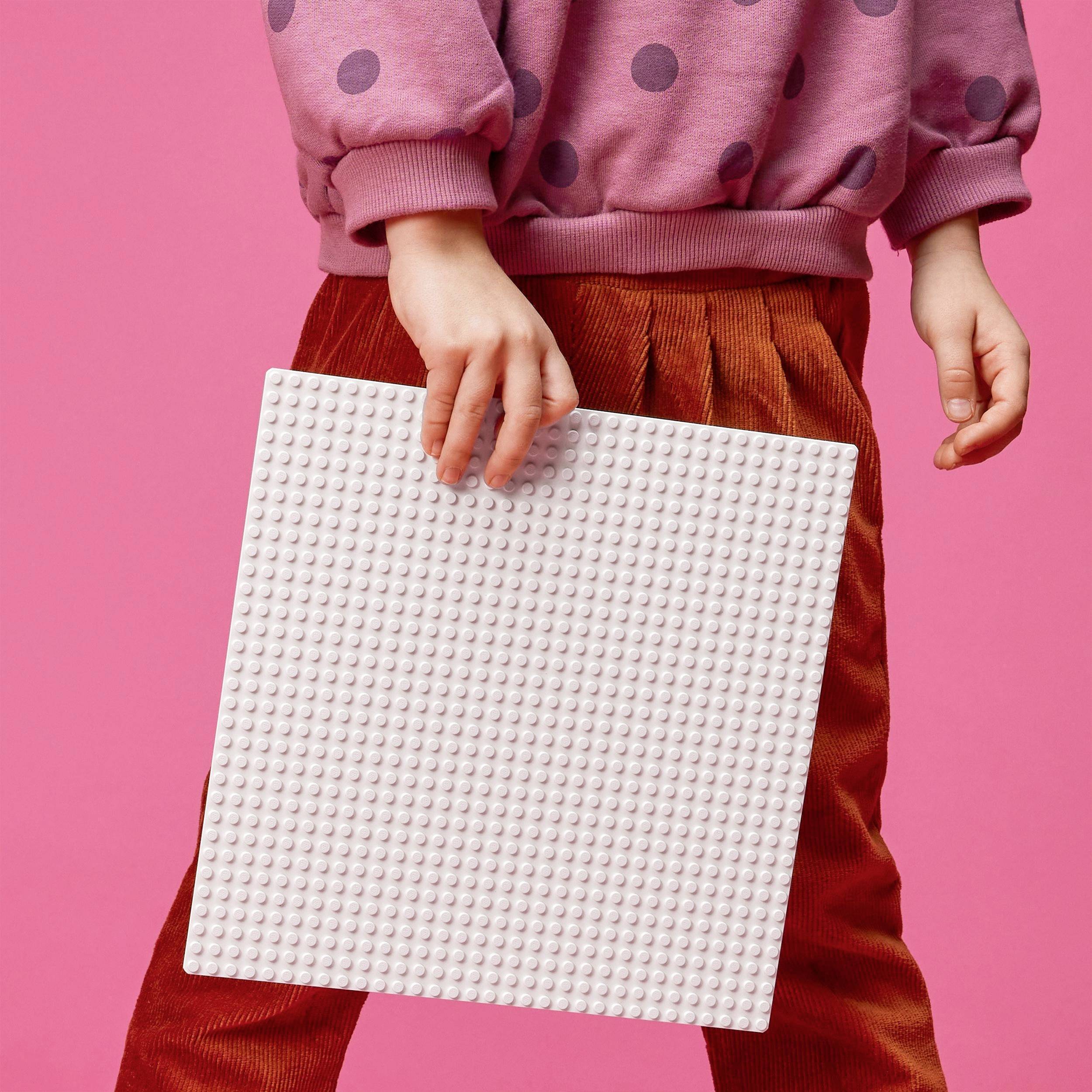 Ett barn i röd klädsel håller ett vitt kvadratiskt byggbräde framför en rosa bakgrund.