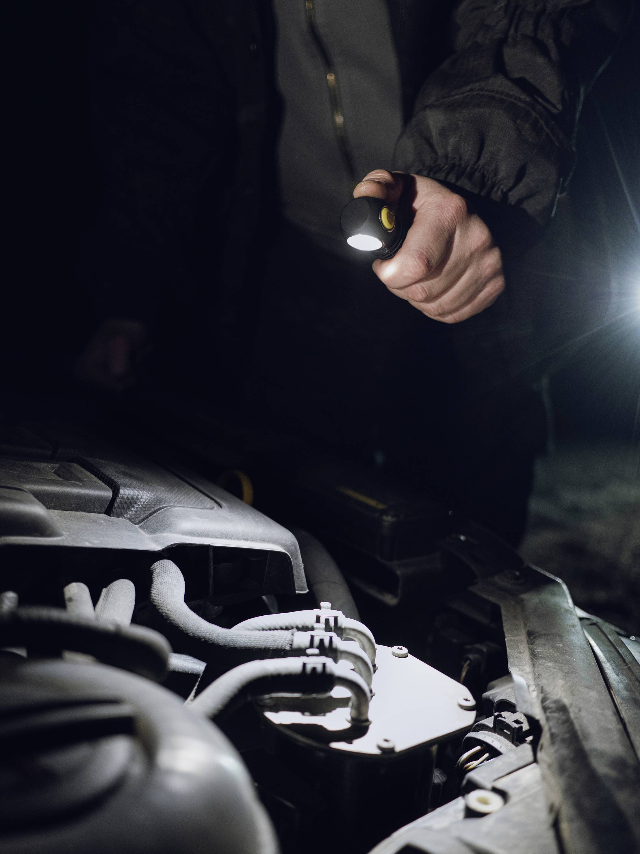 En person lyser med en ficklampa in i motorutrymmet på en bil under natten. Scenen tyder på en bilreparation.