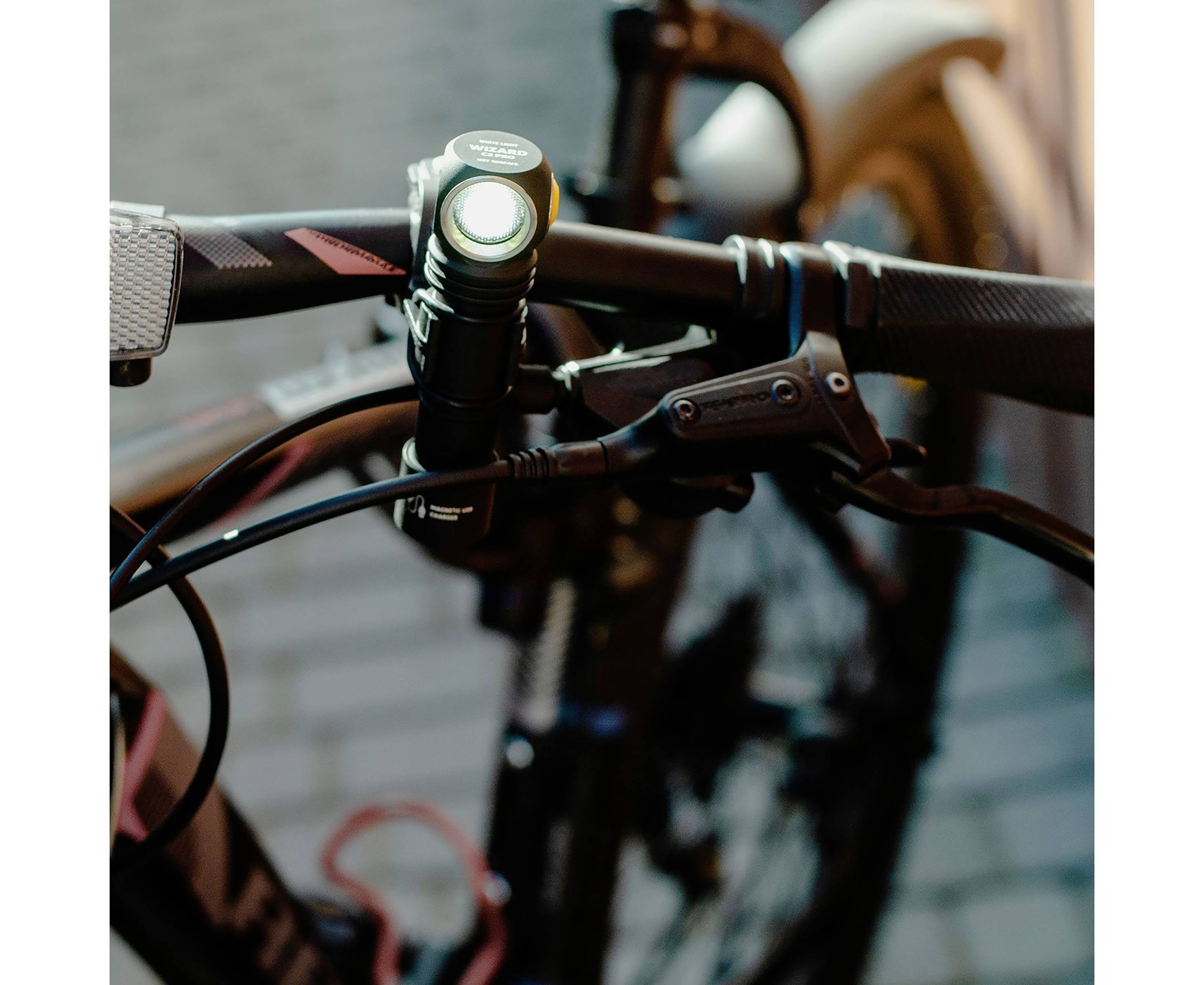 Närbild av en cykelstyre med en monterad ficklampa som är tänd. I bakgrunden suddiga gatstenar.