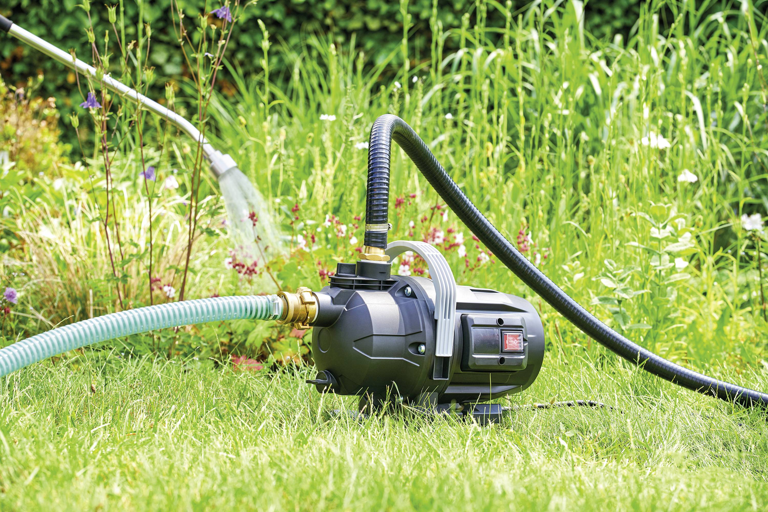 En svart vattenpump på grönt gräs pumpar vatten i en trädgårdsslang som vattnar växter i en blommande trädgård.