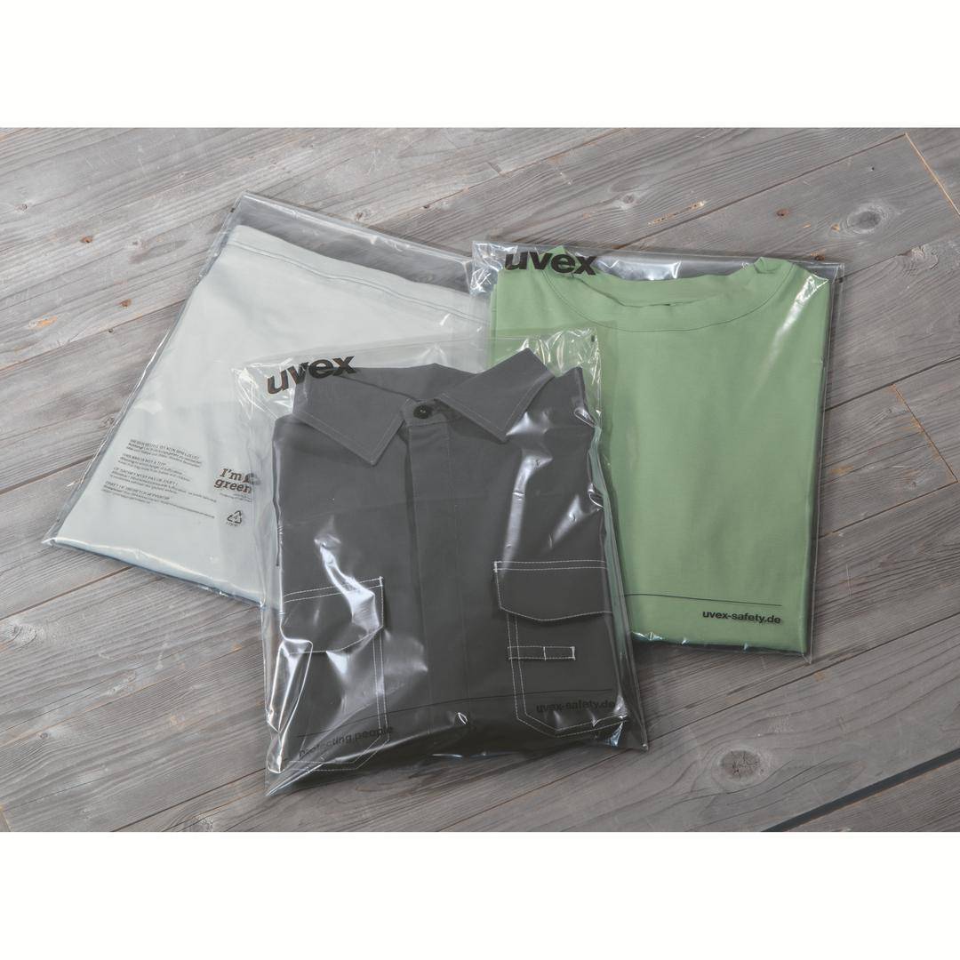 Tre klädesplagg förpackade i plast på ett trägolv, däribland en grå skjorta och en grön t-shirt med logotypen 'uvex'.