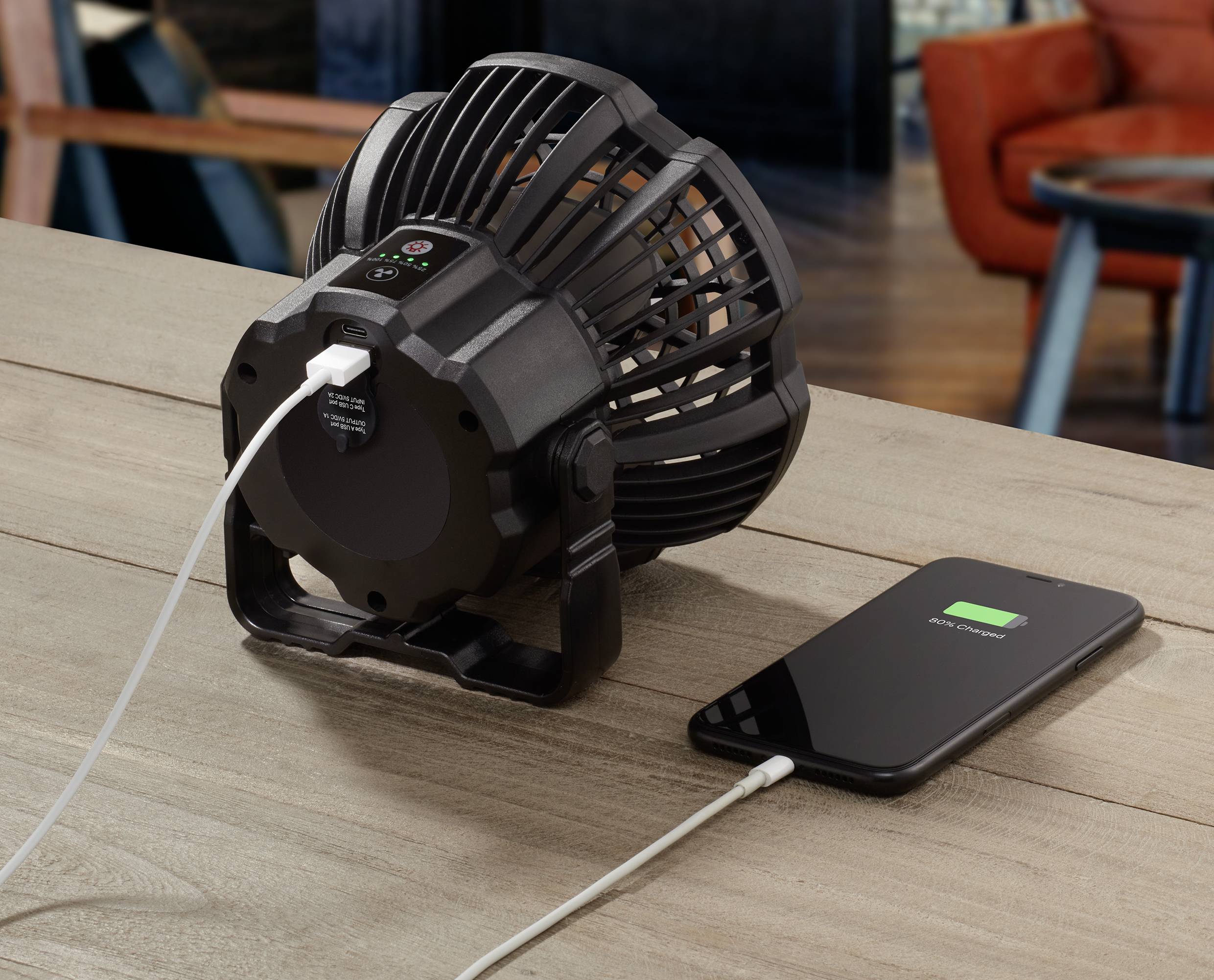 Liten fläkt med USB-anslutning laddar en smartphones på ett träbord. Bakgrunden visar suddiga möbler.