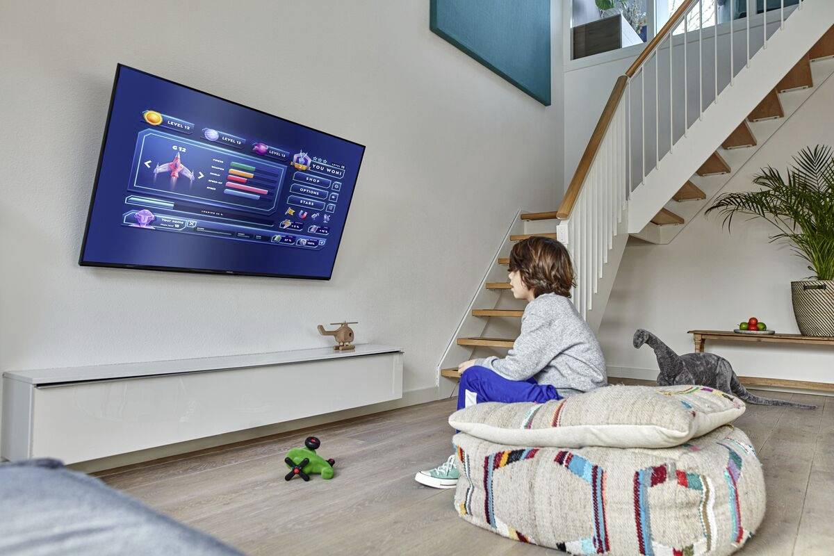 En pojke sitter på kuddar och tittar på en tv med en videospelskärm. En trappa och växter syns i bakgrunden.