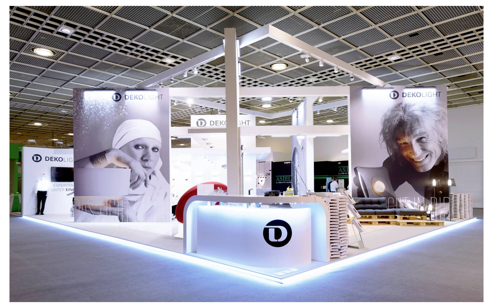 Modern DEKOLIGHT-mässmonter med upplyst disk, varumärkespaneler och stora porträttgrafiker under ett rutnätstak.