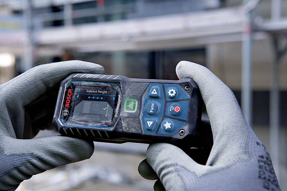 En person med grå arbetshandskar håller ett digitalt Bosch-lasermätinstrument som används för att mäta avstånd.