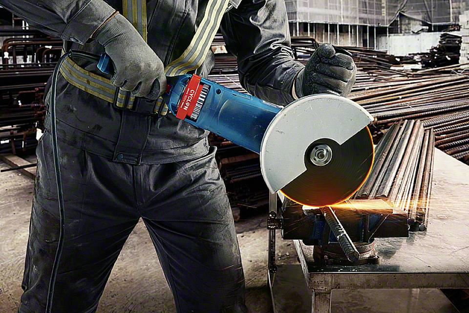 En arbetare i skyddsutrustning klipper metallstänger med en vinkelslip, gnistor flyger around. Tunga maskiner i bakgrunden.