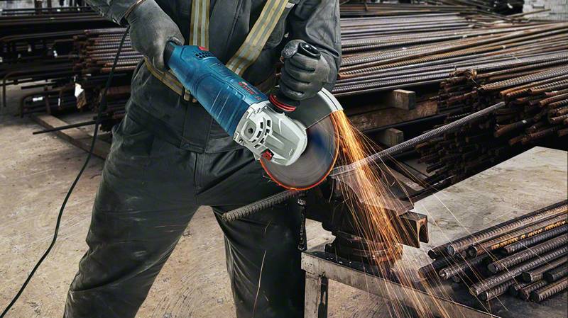Arbetare sågar metallstång med vinkelslip, gnistor flyger. I bakgrunden ligger ytterligare metallstänger staplade.