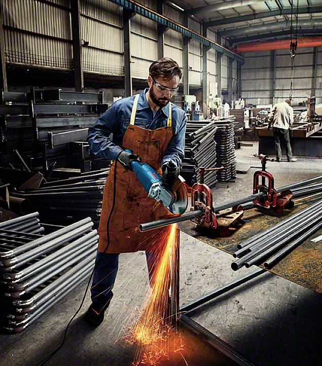 En man i skyddskläder slipar metall i en verkstad och skapar gnistor. I bakgrunden syns en industriell miljö.