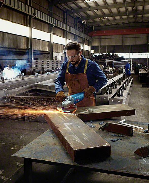 En man i en verkstad slipar metall med en vinkelslip, gnistor spritter. Omgivningen visar industriell metallbearbetning.