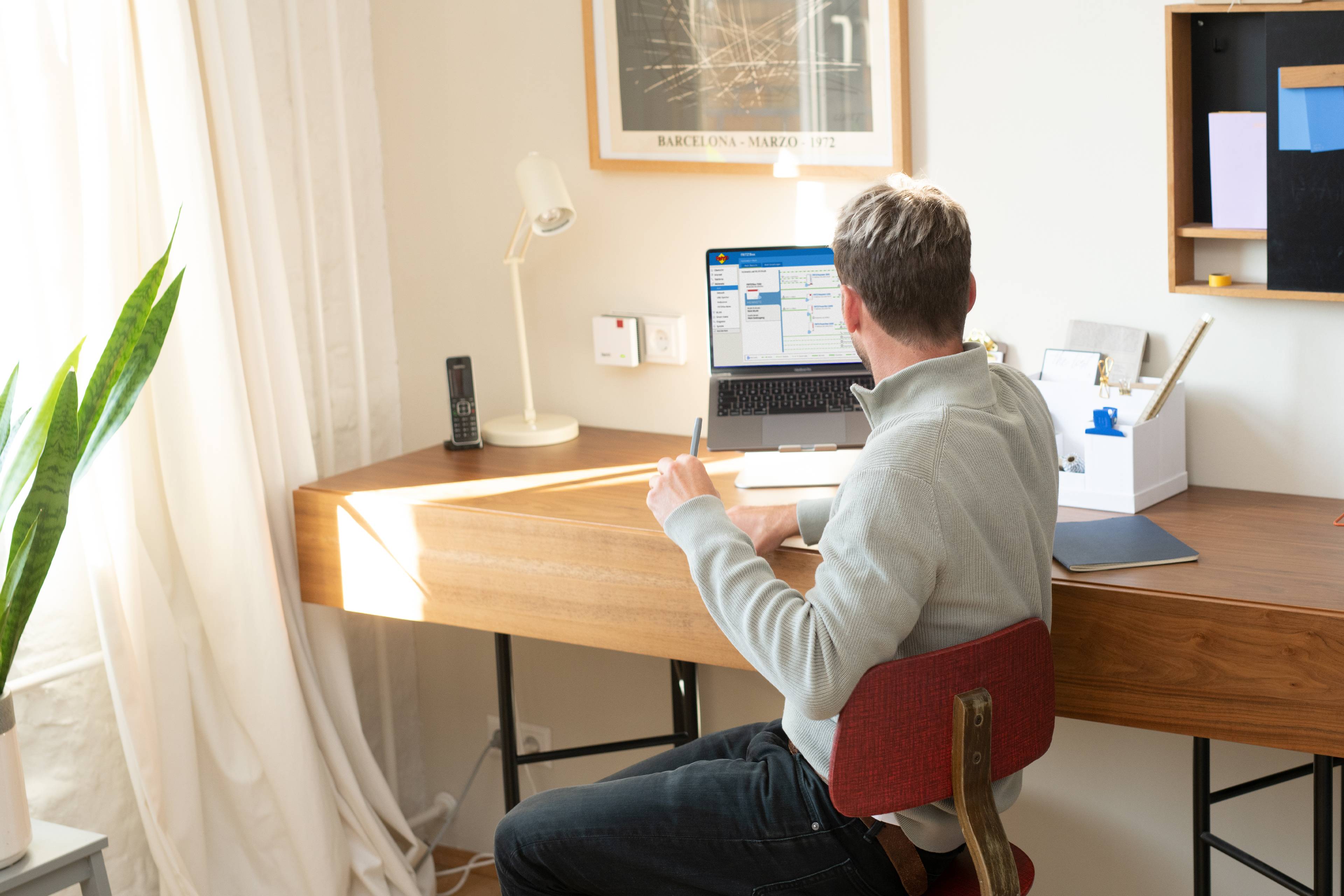 En man sitter vid ett skrivbord och arbetar på en bärbar dator. Rummet är ljust upplyst, med växter och kontorsmaterial runt omkring sig.