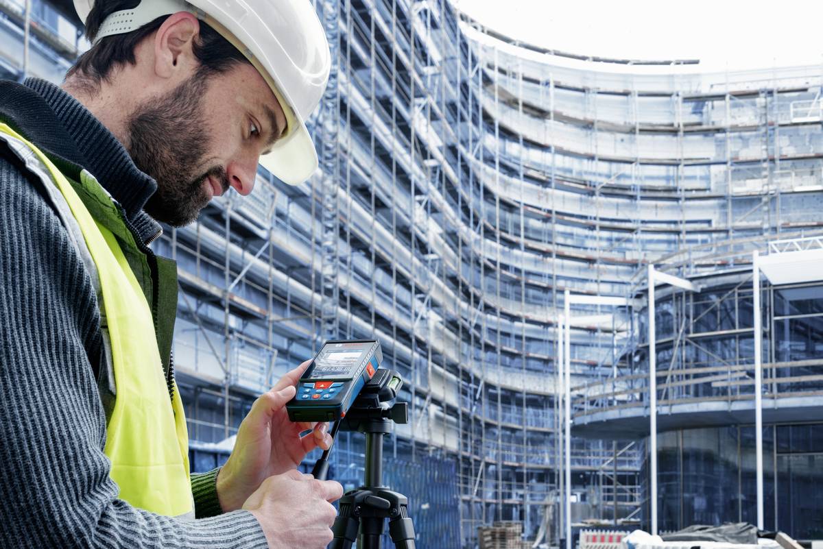 En byggnadsarbetare med skyddshjälm och varselväst använder ett lasermätinstrument på en byggarbetsplats framför en ställningsförsedd byggnad.