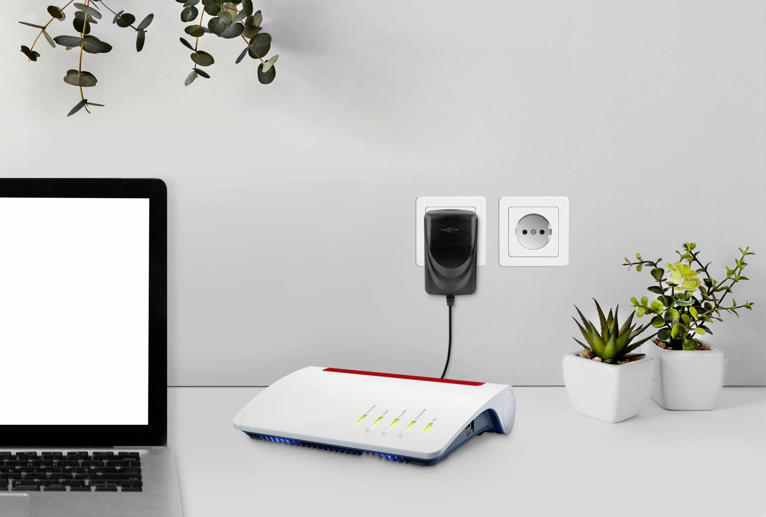Bärbar dator och router på ett skrivbord, omgivna av växter. Routern är ansluten till ett eluttag och visar aktiva lampor.